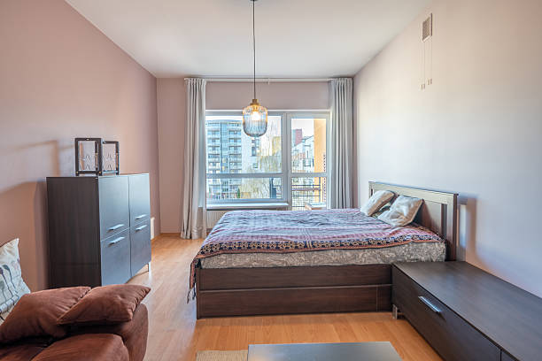Brightly lit modern bedroom featuring a large bed, wooden furniture, soft textiles, and a view of an urban landscape through large windows